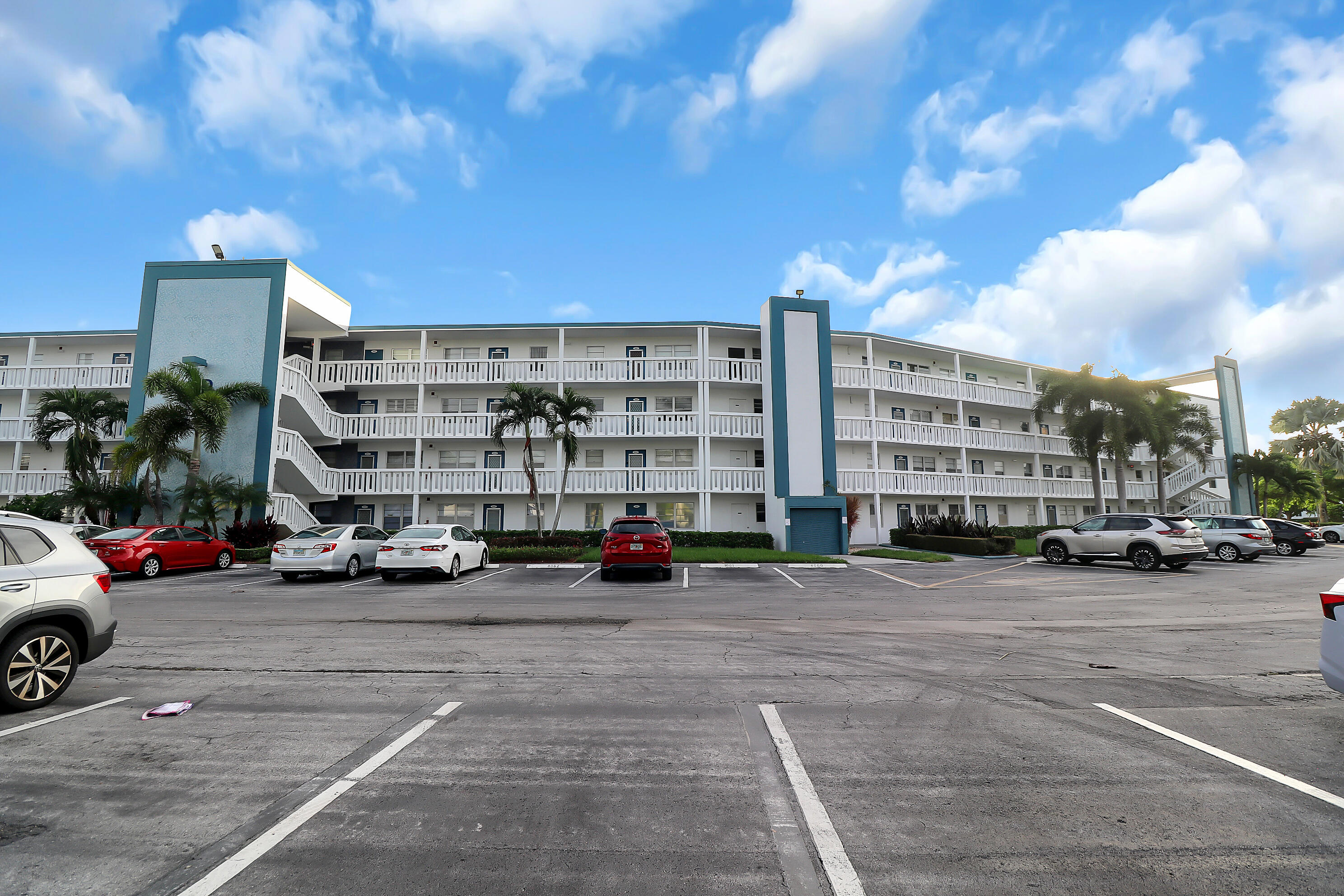 Multi-story condominium building with white balconies, teal accents, palm trees, and a front parking lot.