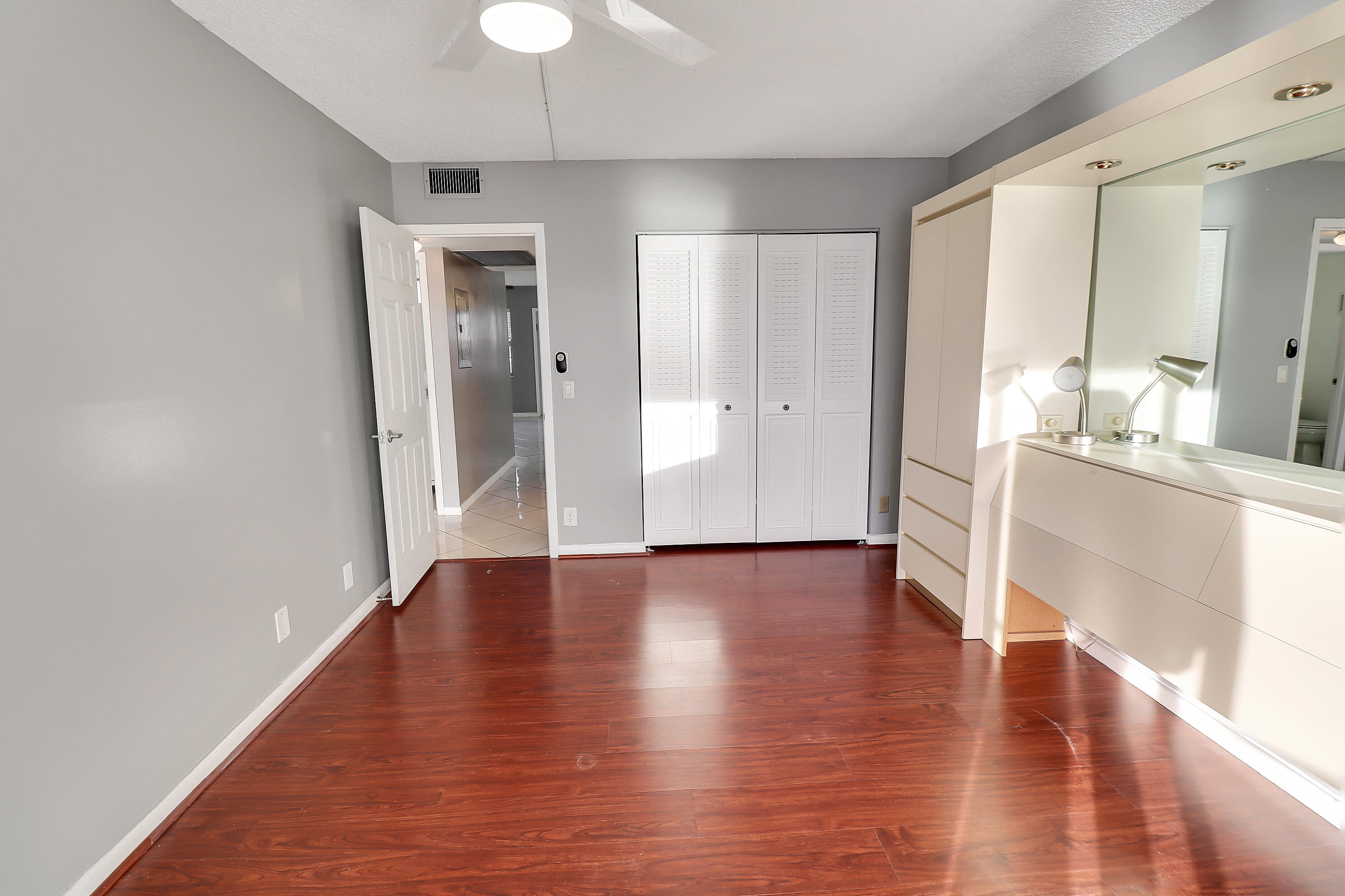 Empty bedroom with reddish wood floors, a mirrored vanity, and white louvered closet doors.