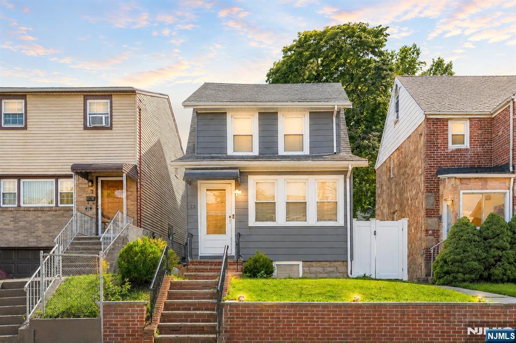 Gray two-story house with white trim, front lawn, brick retaining wall, and neighboring homes.