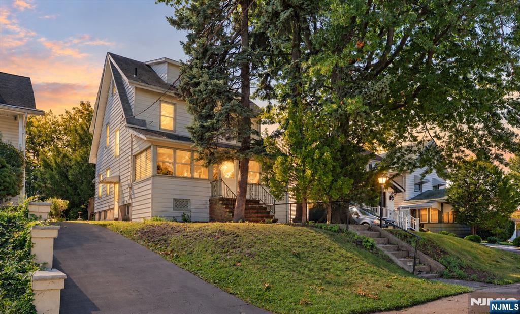 Two-story light-colored home with glowing windows, a sloped lawn, and mature trees at sunset.