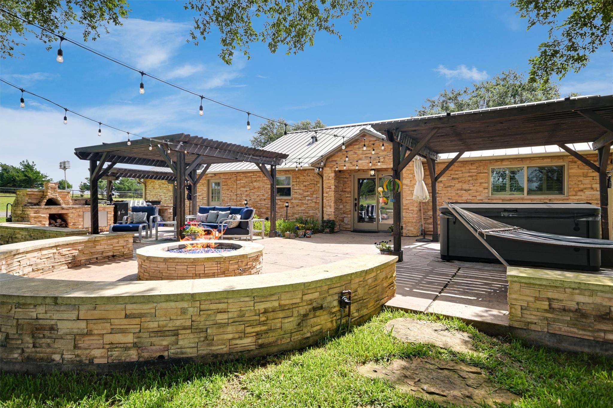 Stone home patio with pergola seating, a circular fire pit, string lights, and a hot tub.
