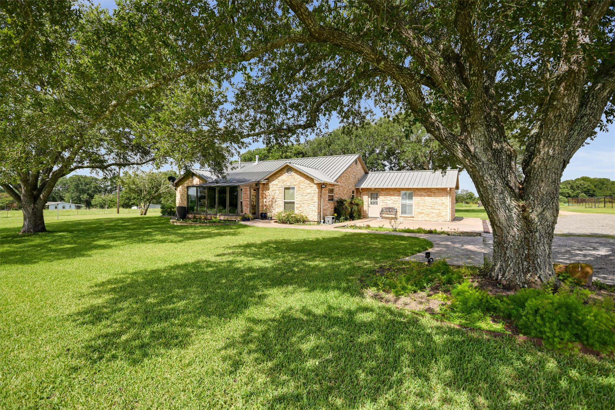 Stone house with a metal roof framed by mature trees, with a broad green lawn and paved patio.