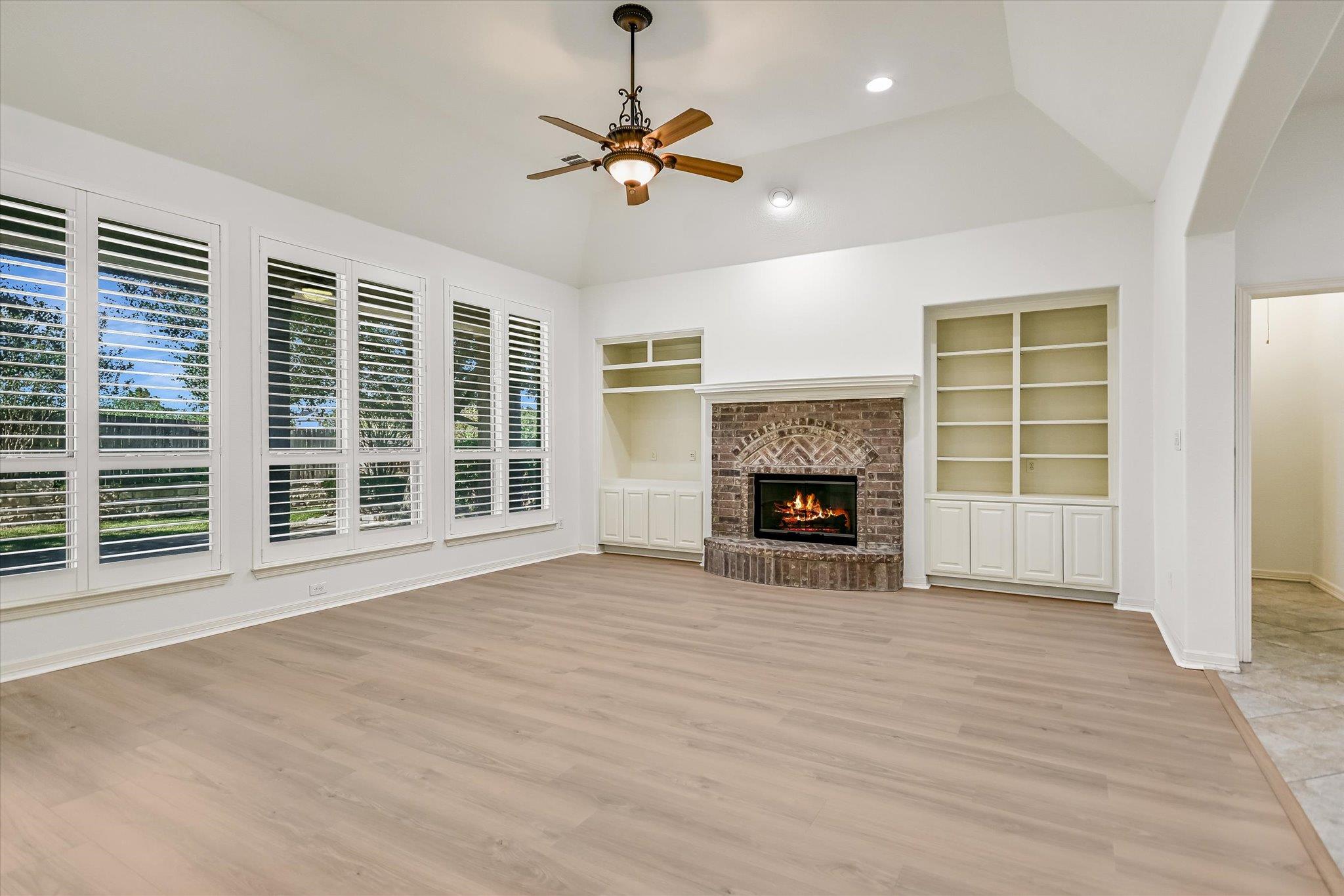 Empty living room with a brick fireplace, built-in shelving, and plantation shutters.