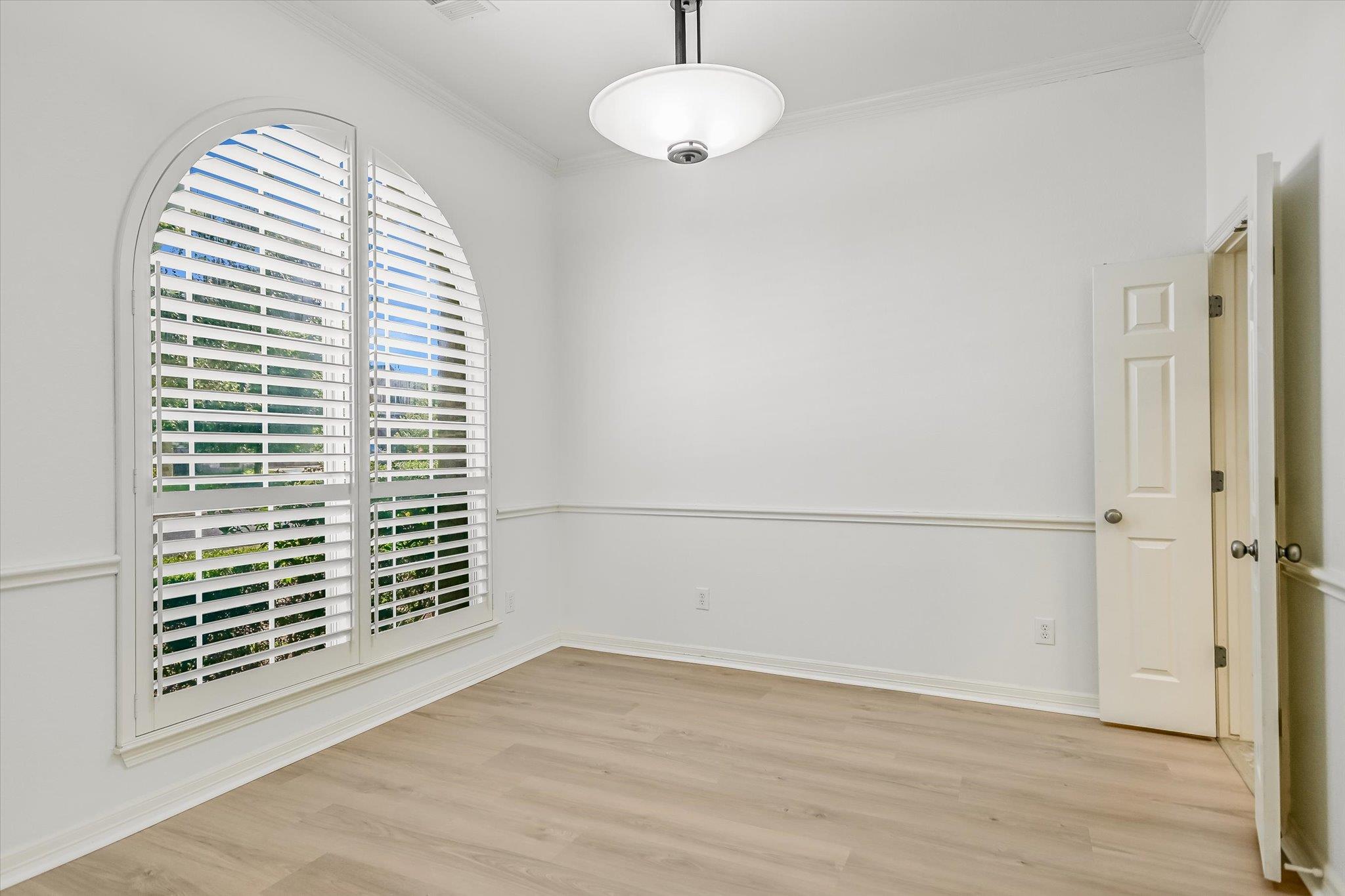 Empty room with arched plantation-shutter windows, light wood flooring, and a ceiling light.