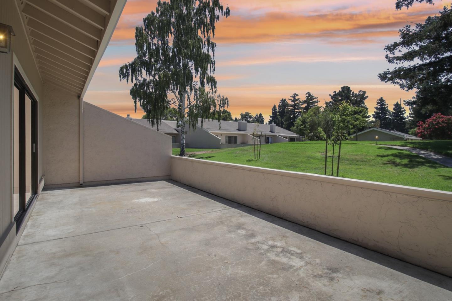Empty concrete patio overlooking a green lawn and trees beneath a pink-orange sunset sky.