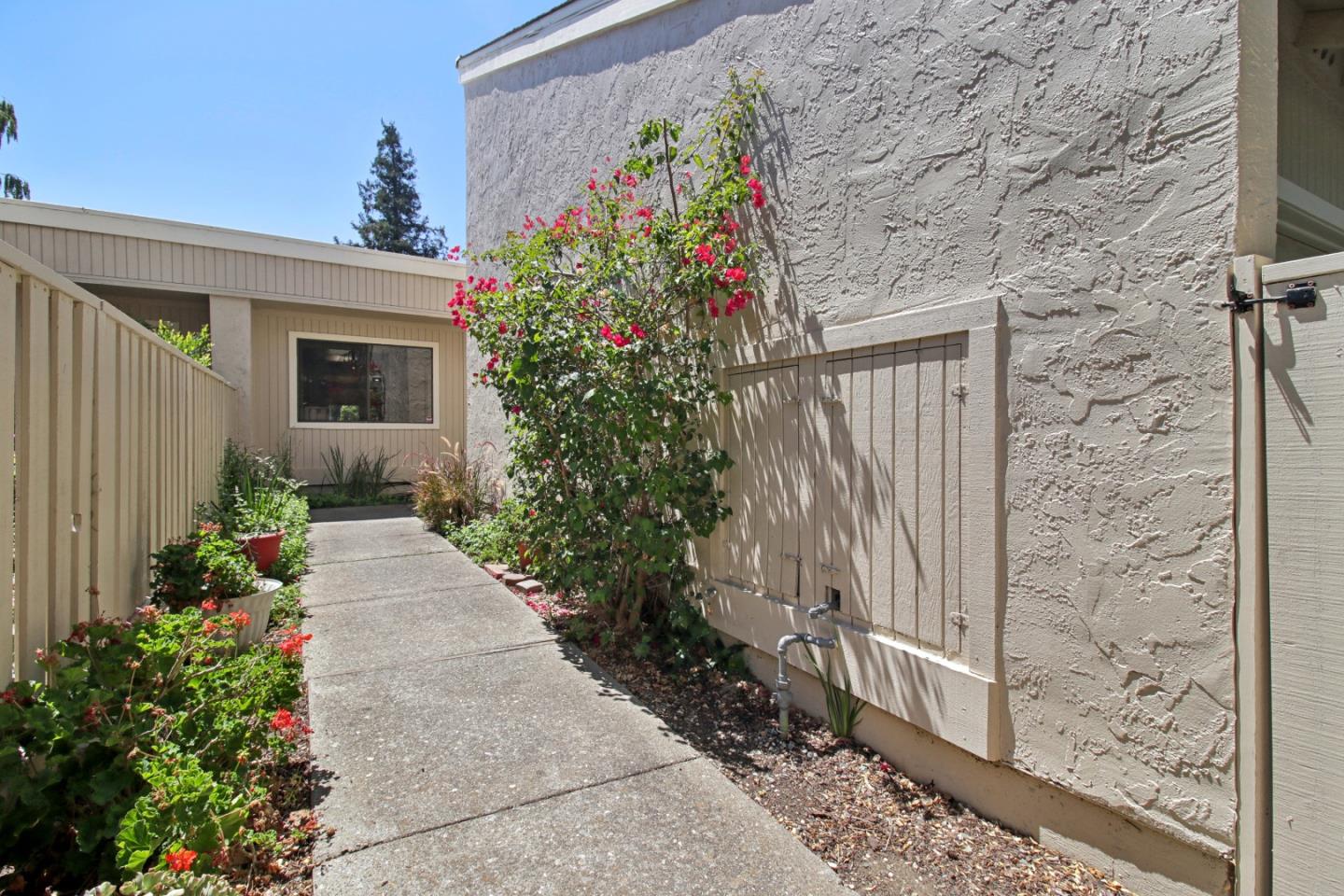 Concrete walkway beside a stucco home, bordered by a fence and flowering plants.