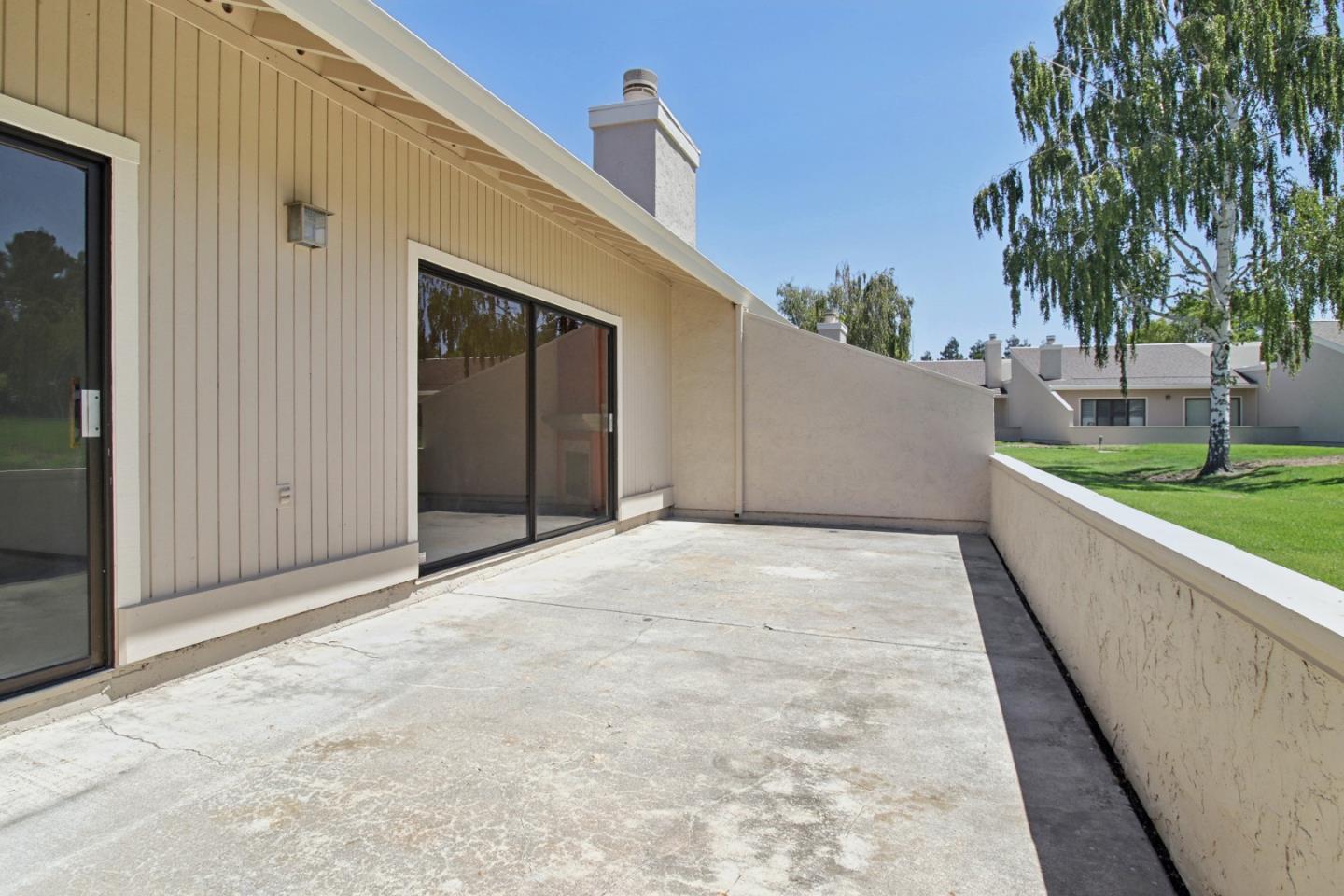 Concrete patio beside a beige home, bordered by a low wall overlooking a lawn and trees.