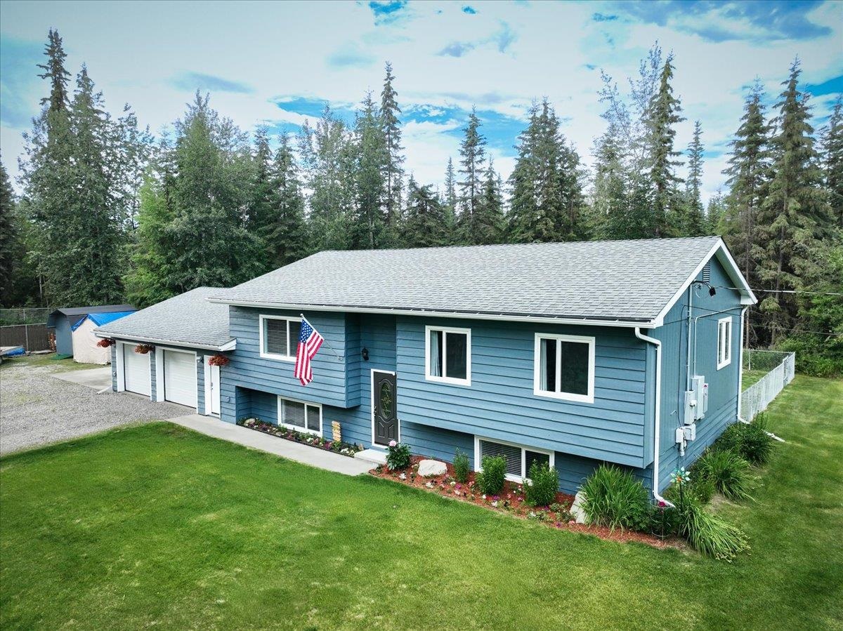 Blue-sided home with an attached garage, green lawn, and evergreen forest behind it.