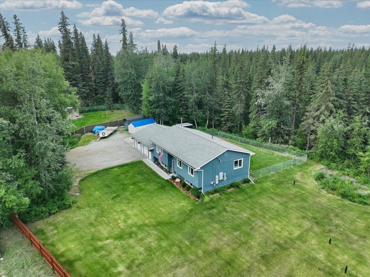 Aerial view of a blue home on a fenced grassy lot surrounded by forest.