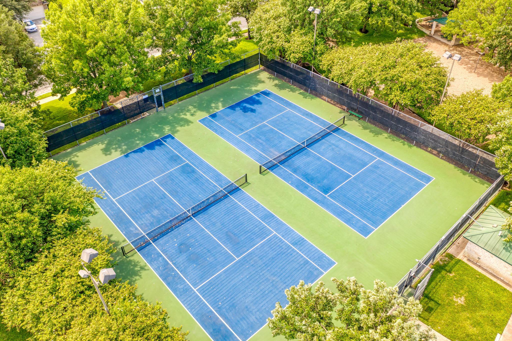 Two blue tennis courts surrounded by trees and fencing, seen from above.