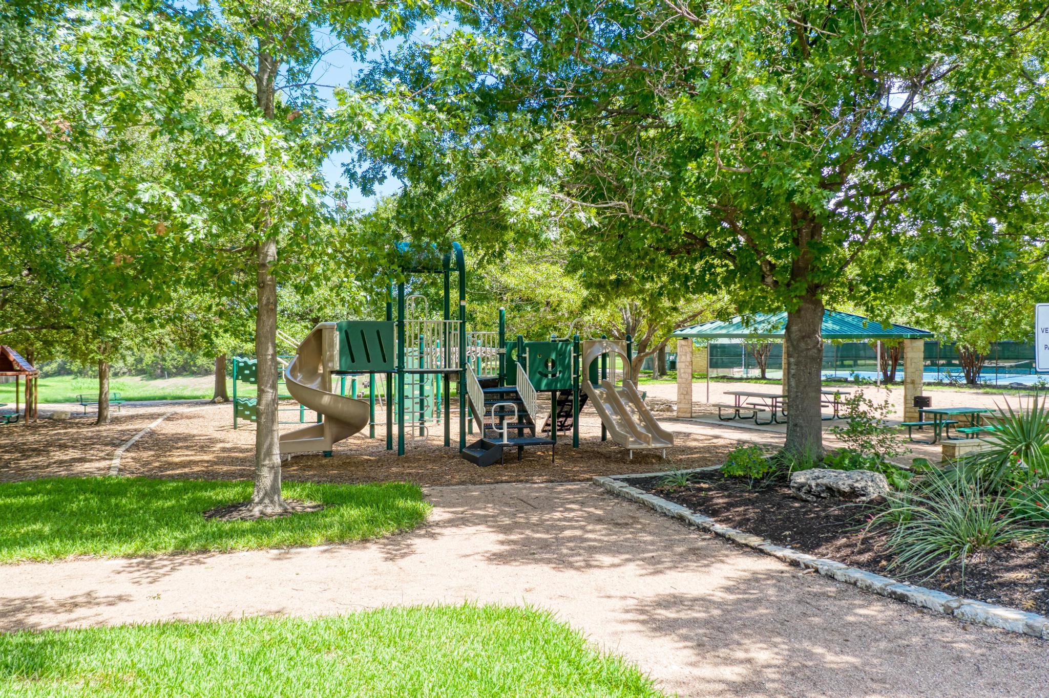 Community playground with slides beneath mature trees, beside a covered pavilion and fenced pool.
