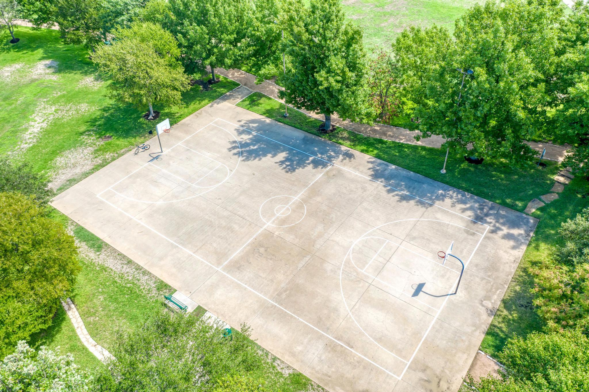 Aerial view of a basketball court with two hoops, bordered by mature trees and walking paths.