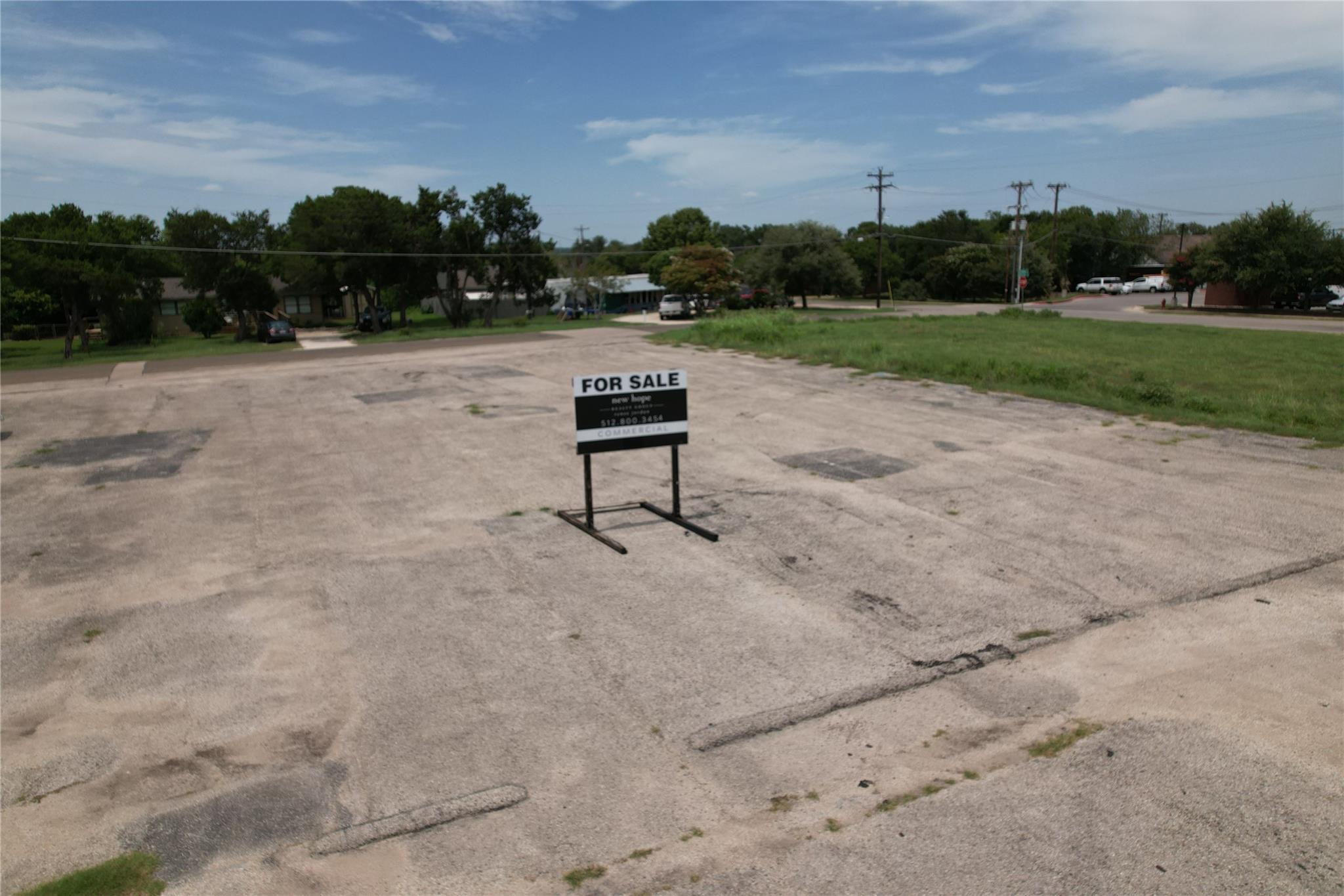 Vacant paved commercial lot with a for-sale sign, trees, and neighboring buildings in the background.