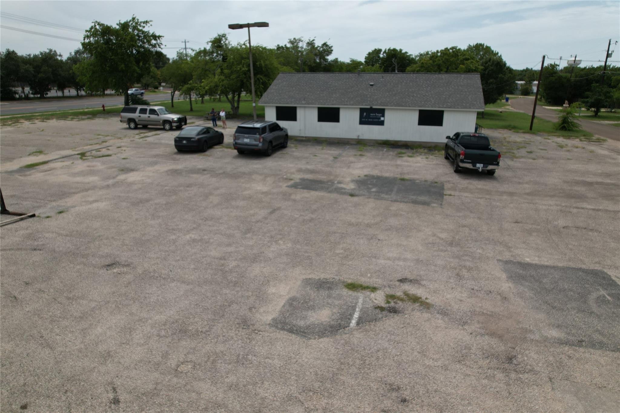 Single-story white commercial building beyond a large paved parking lot with several parked vehicles.