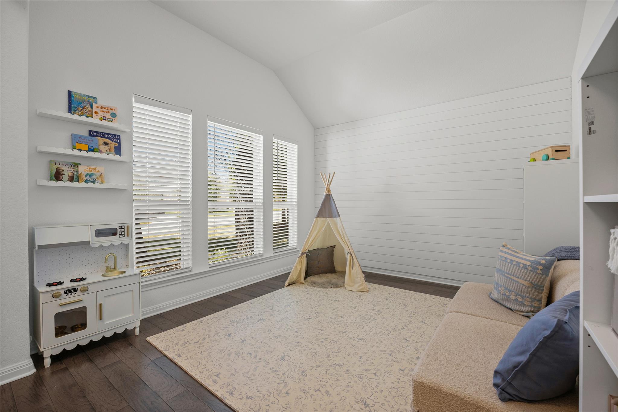 Playroom with a small teepee, toy kitchen, book ledges, and three windows beneath a sloped ceiling.
