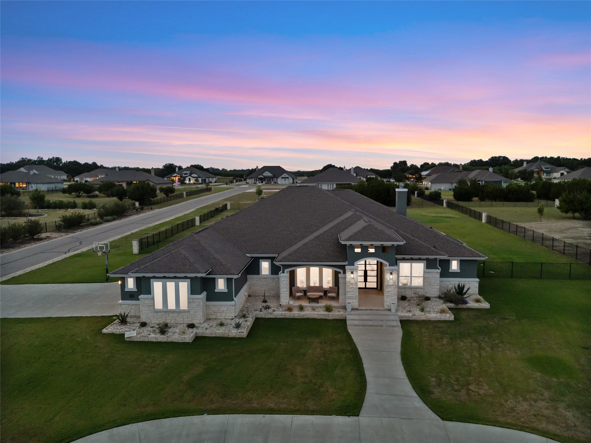 Single-story stone-and-siding home with a broad lawn, curved driveway, and neighboring homes at dusk.