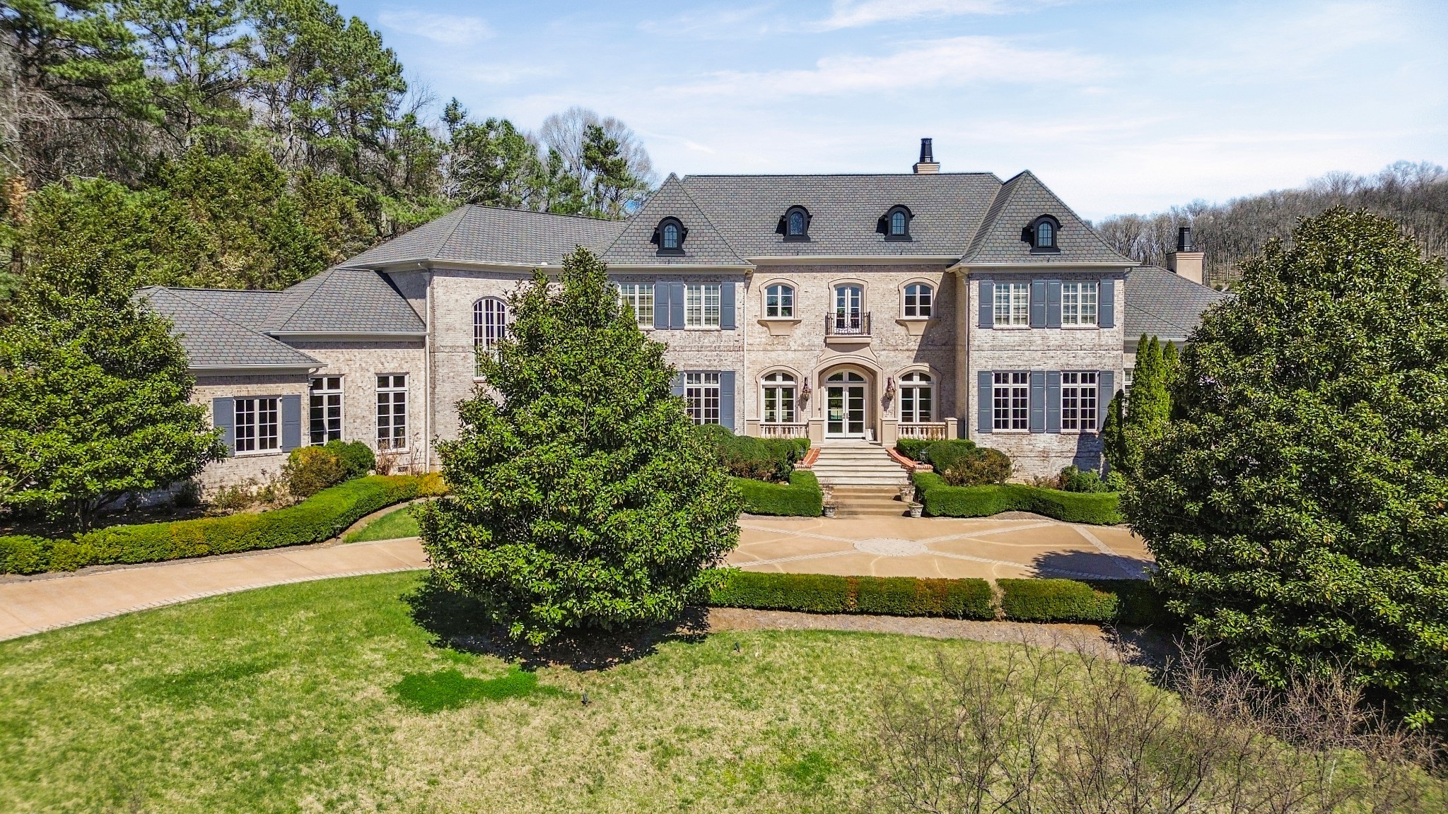 Large light-brick home with gray roofs, blue-gray shutters, and formal landscaped grounds.