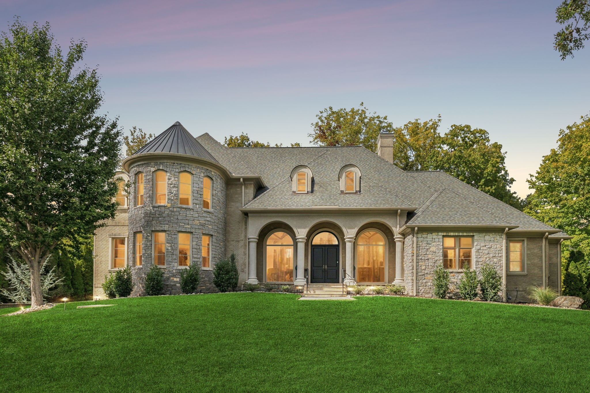 Stone home with a round turret, arched entry, and broad front lawn at dusk.