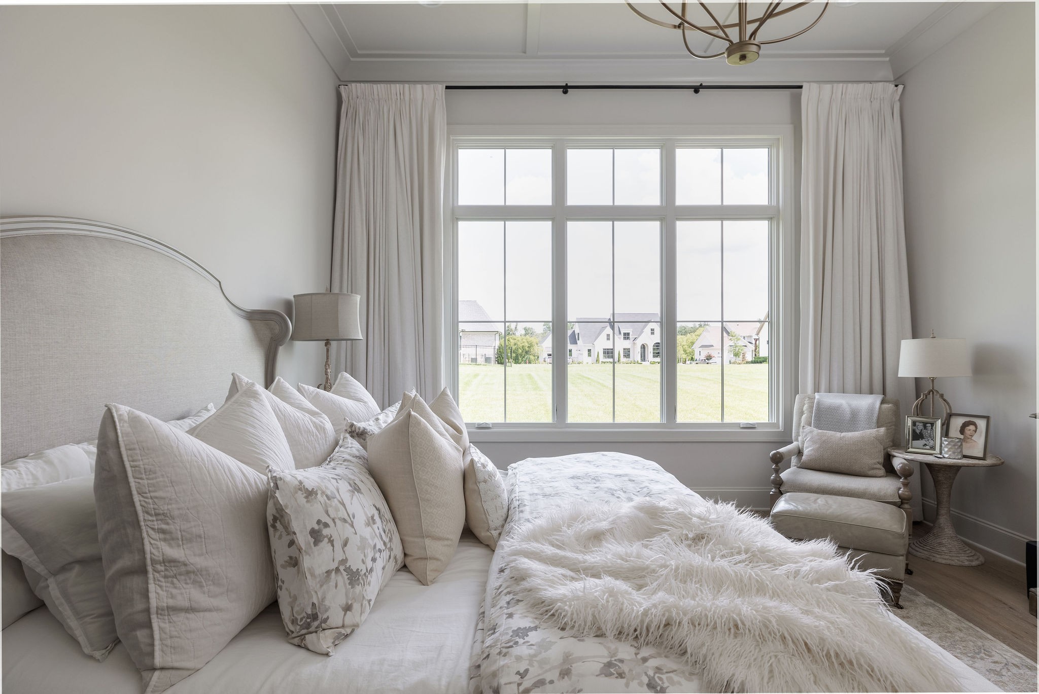 Bright bedroom with a layered white bed, reading chair, and tall windows overlooking a lawn.