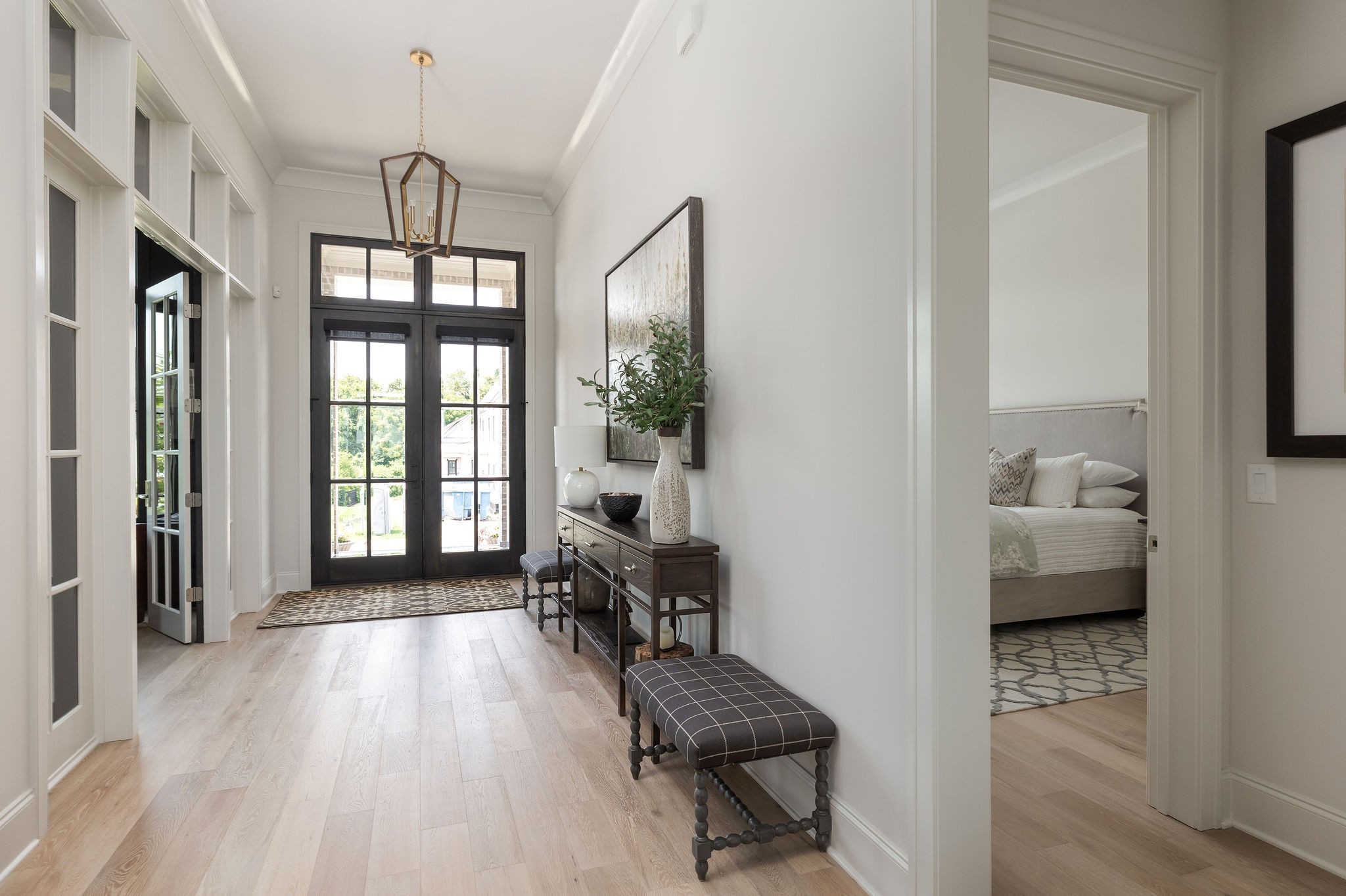 Bright entry hall with pale wood floors, a dark-framed glass front door, and a bedroom visible through the doorway.