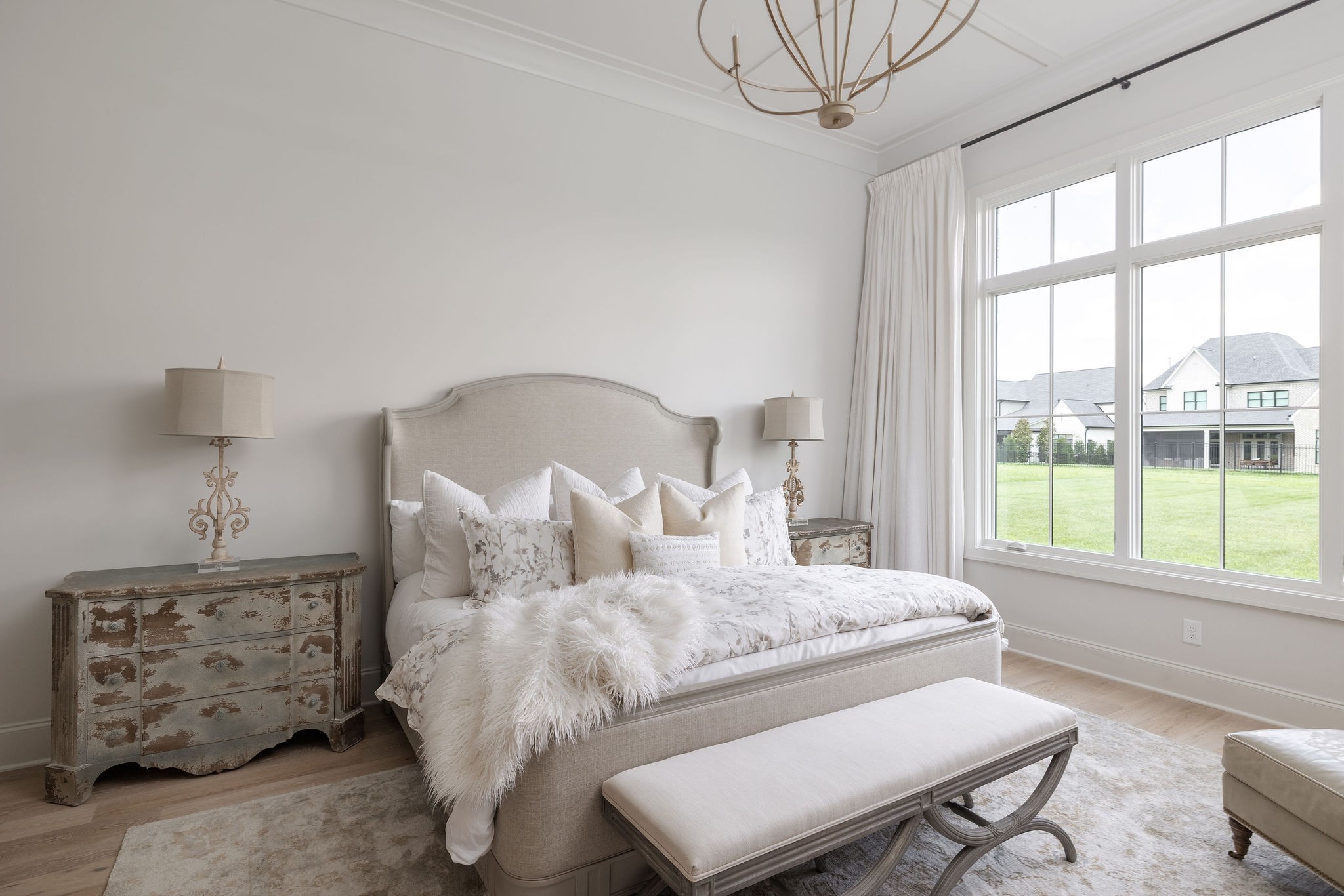 Staged bedroom with an upholstered bed, distressed bedside dresser, and large windows overlooking a lawn.