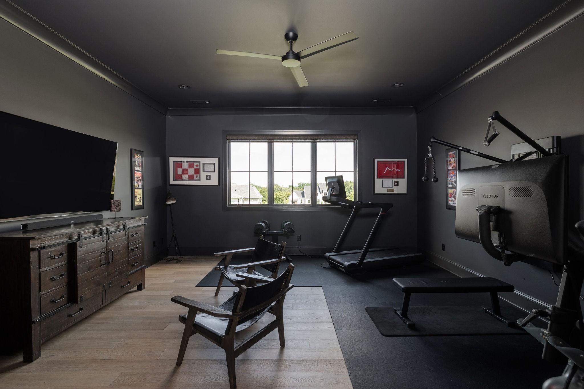 Home gym with a treadmill, Peloton strength machine, weights, and lounge chairs by a large window.
