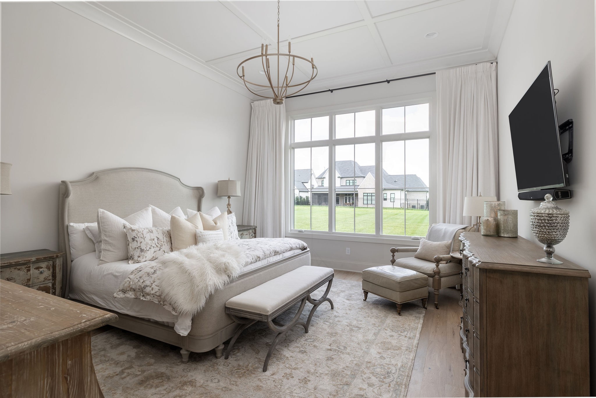 Bedroom with a cream upholstered bed, large curtained window, and armchair beside a dark wood dresser.