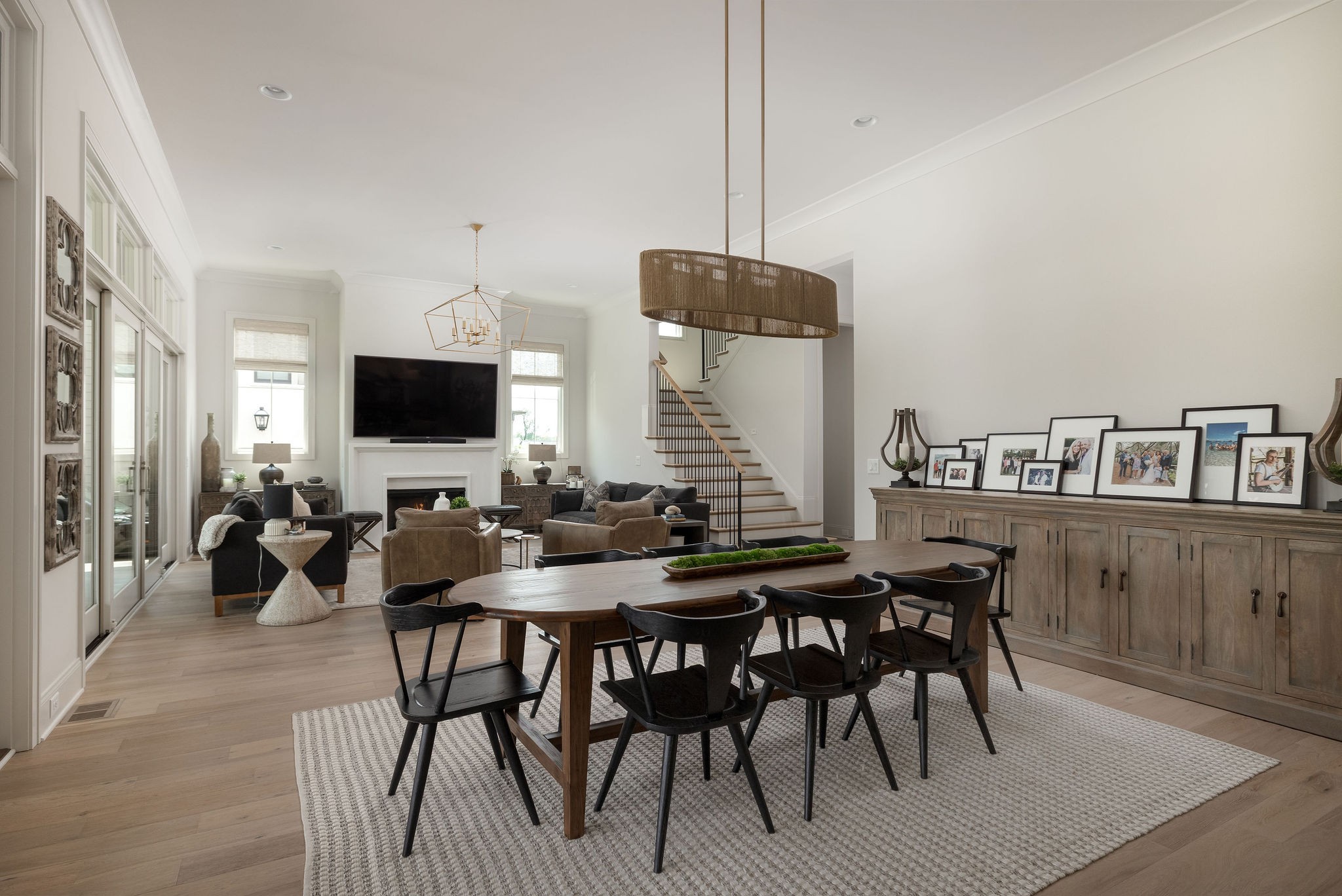 Open-plan dining and living room with a wood table, tall windows, fireplace, and staircase.