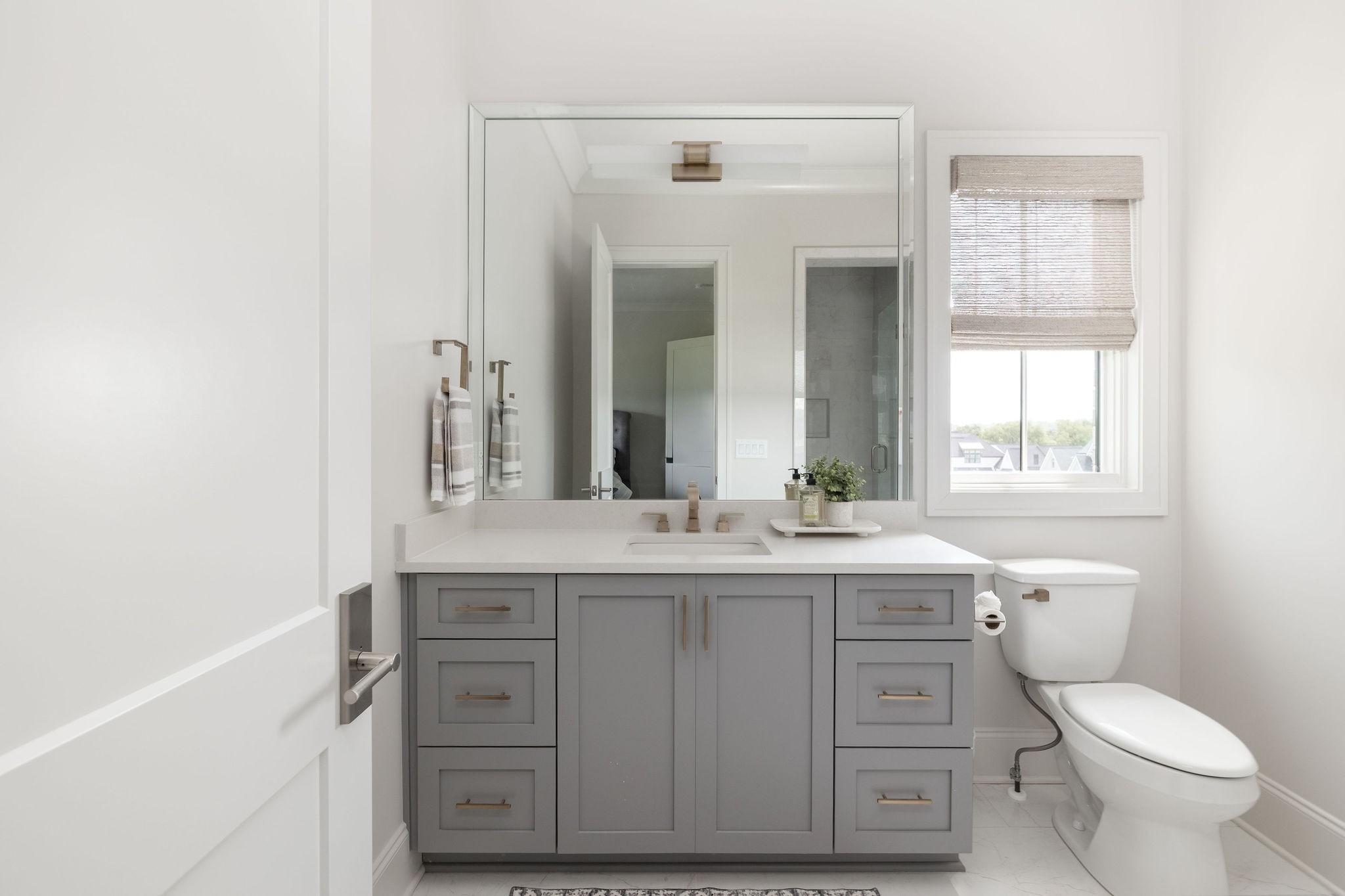 Bright bathroom with a gray vanity, large mirror, and window beside the toilet.
