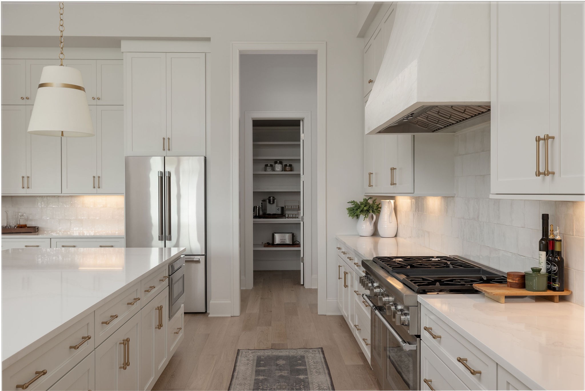 White kitchen with a large island, professional gas range, stainless refrigerator, and open walk-in pantry.