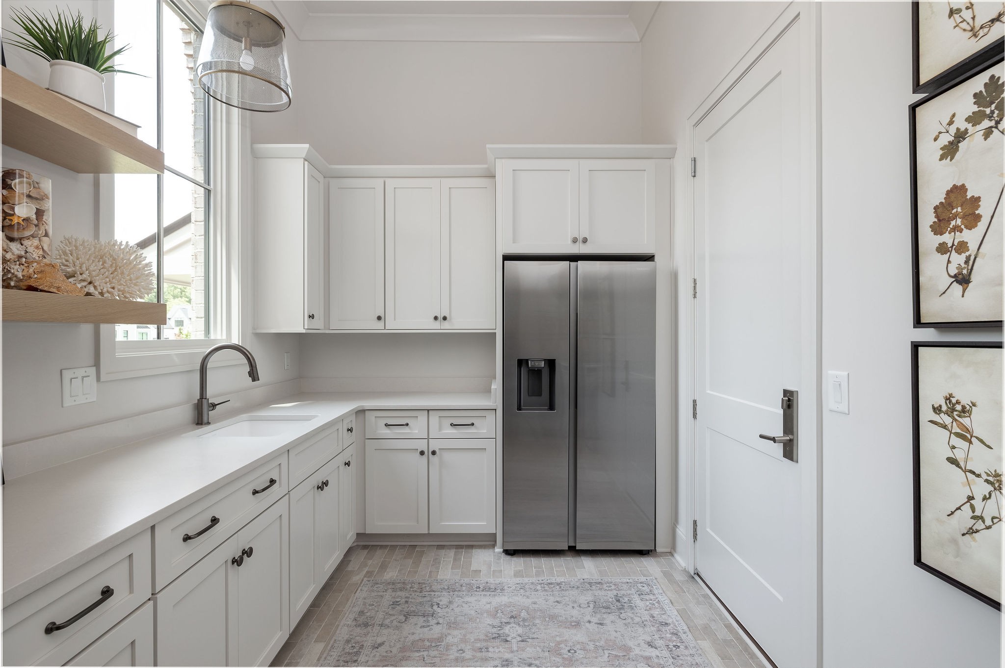 Bright white kitchen with shaker cabinets, stainless refrigerator, and sink beneath a tall window.