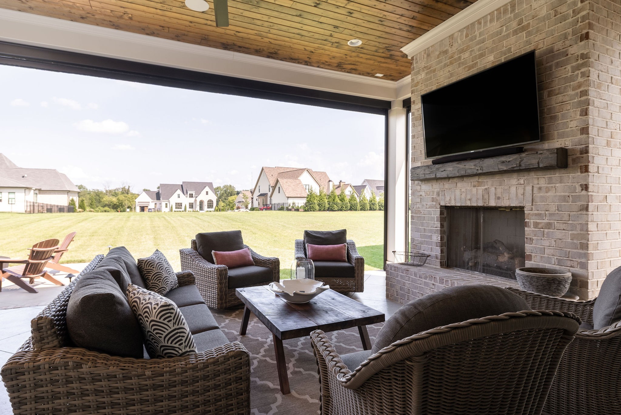 Covered patio with wicker seating, a brick fireplace, and a view across the lawn to neighboring homes.