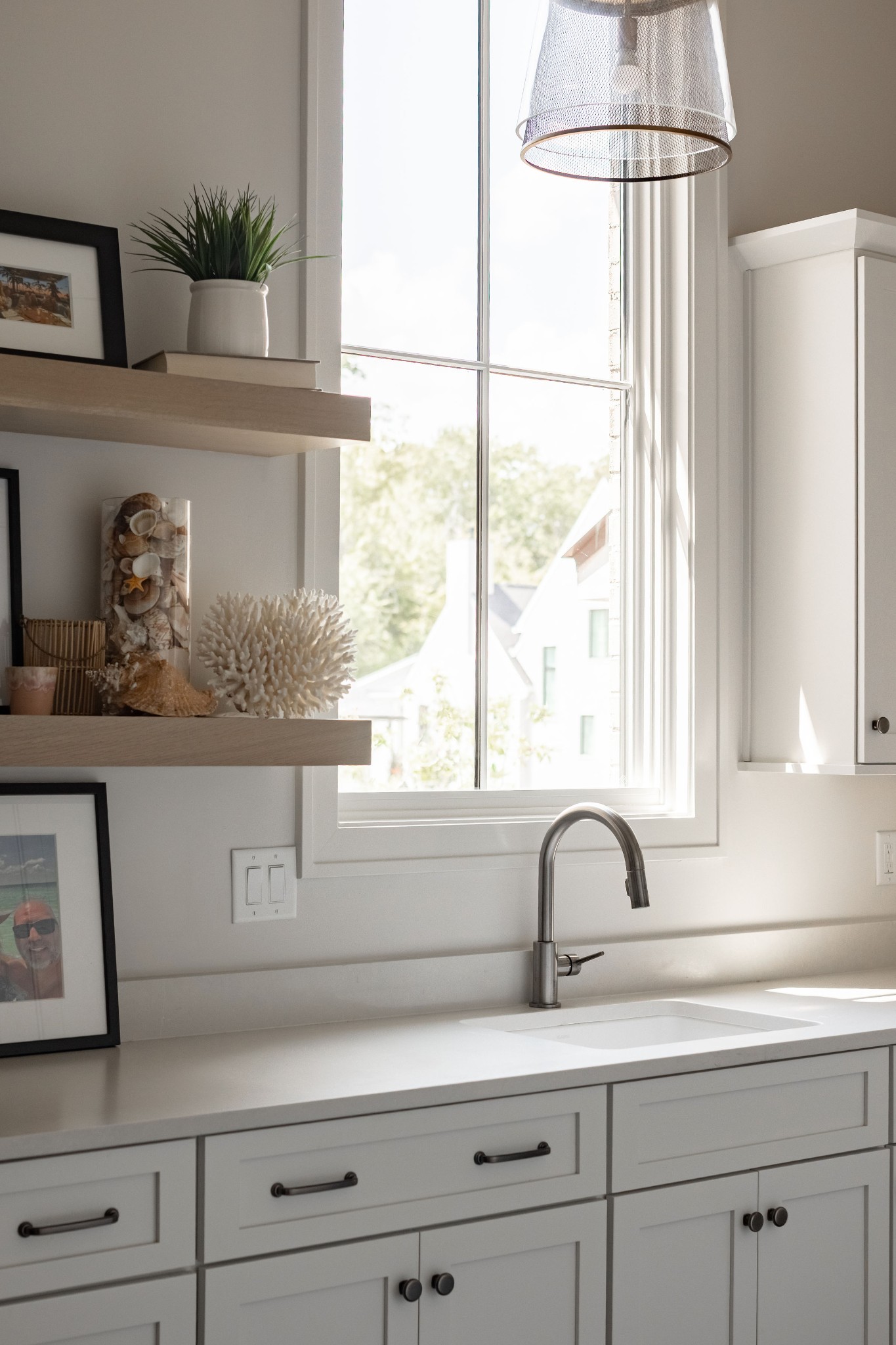 Bright white kitchen sink beneath a large window, with open shelves displaying plants and seashells.