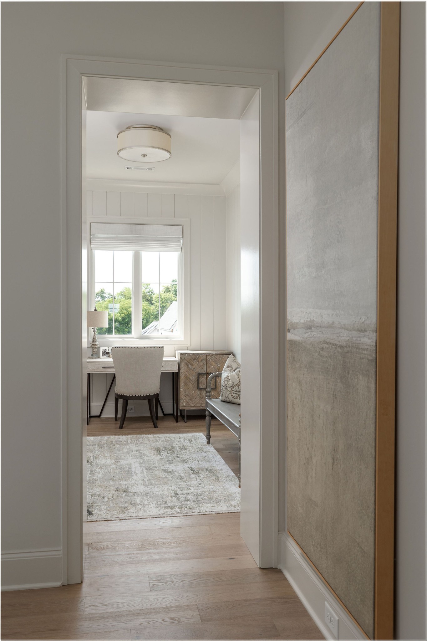 Furnished home office with a desk and chair by a bright window, viewed through a doorway.