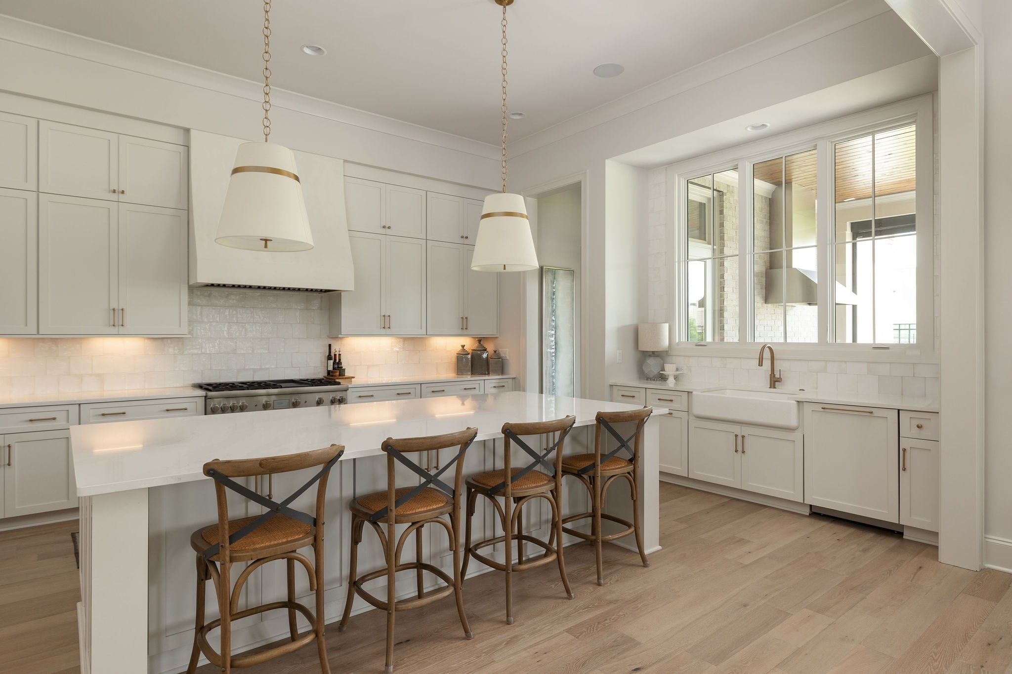 Bright white kitchen with a large island, four wooden stools, and a farmhouse sink beneath a wide window.