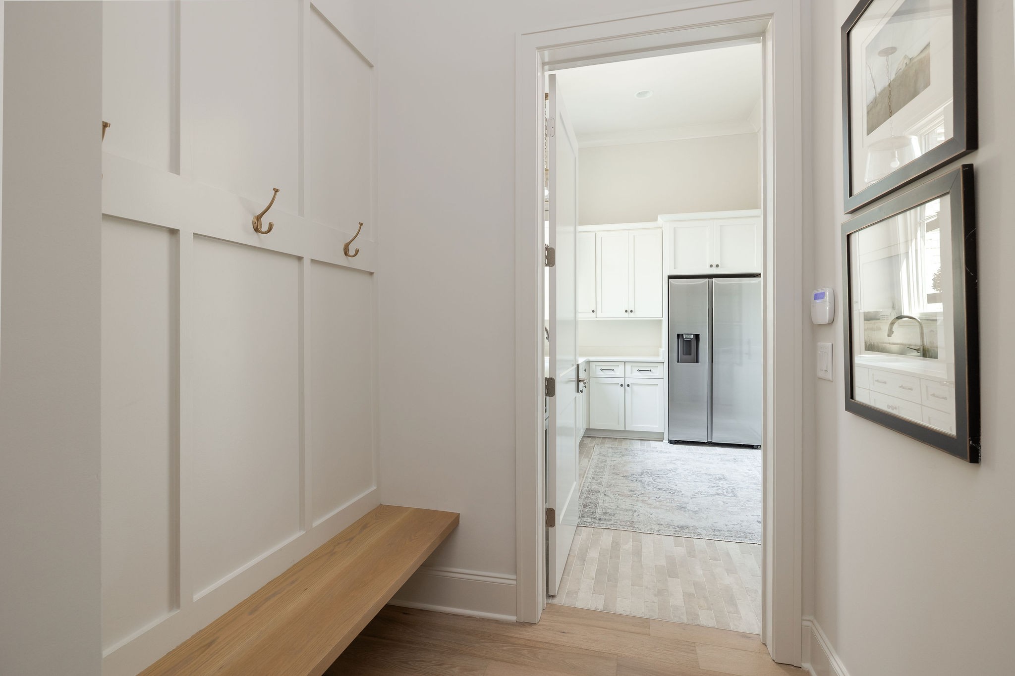 Mudroom with a built-in wood bench and brass hooks, opening onto a bright kitchen with a stainless steel refrigerator.