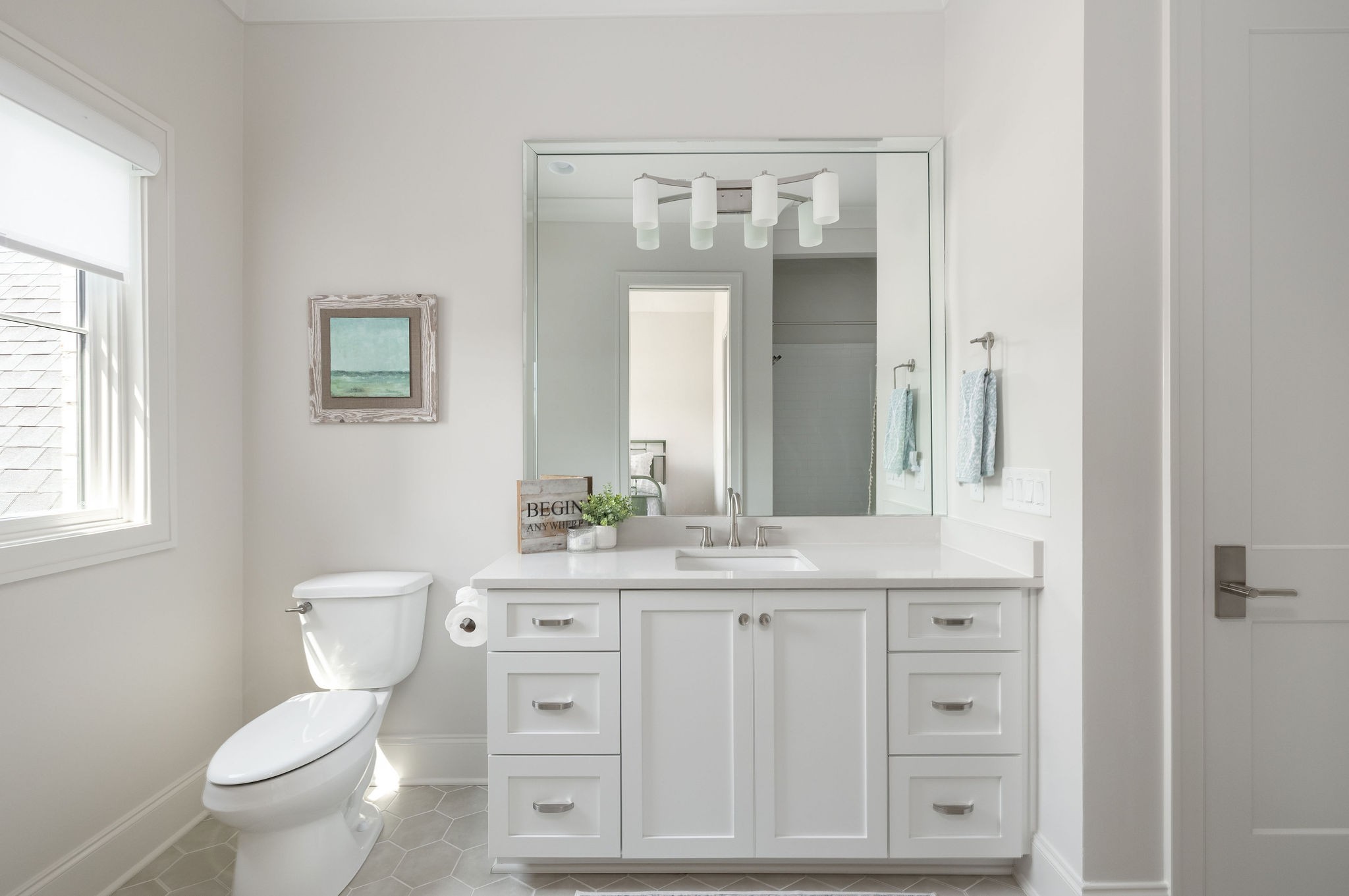 Bright bathroom with a white mirrored vanity, toilet, gray hexagonal tile floor, and coastal artwork.