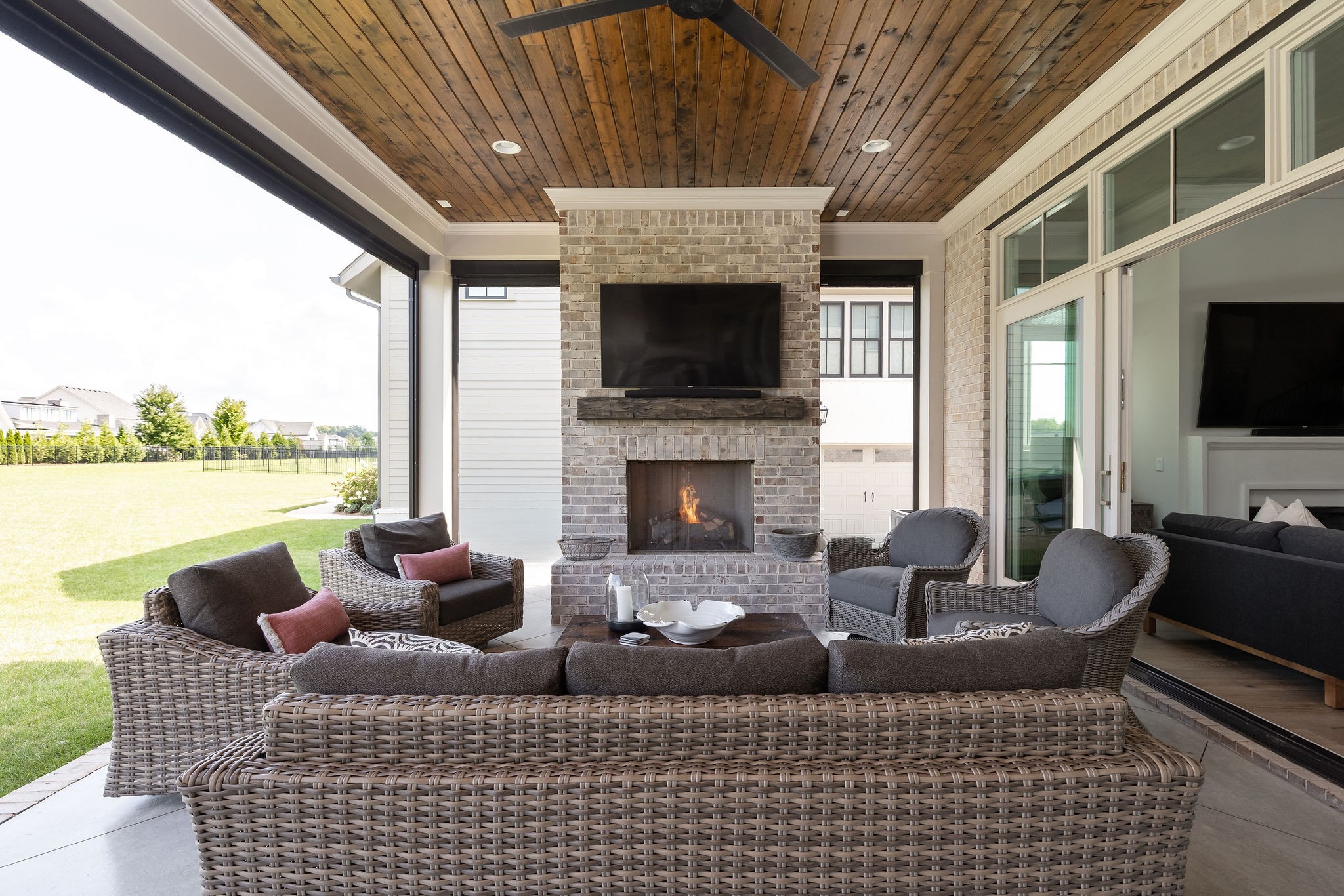 Covered patio with wicker seating, a brick fireplace, and a wood-plank ceiling overlooking the lawn.