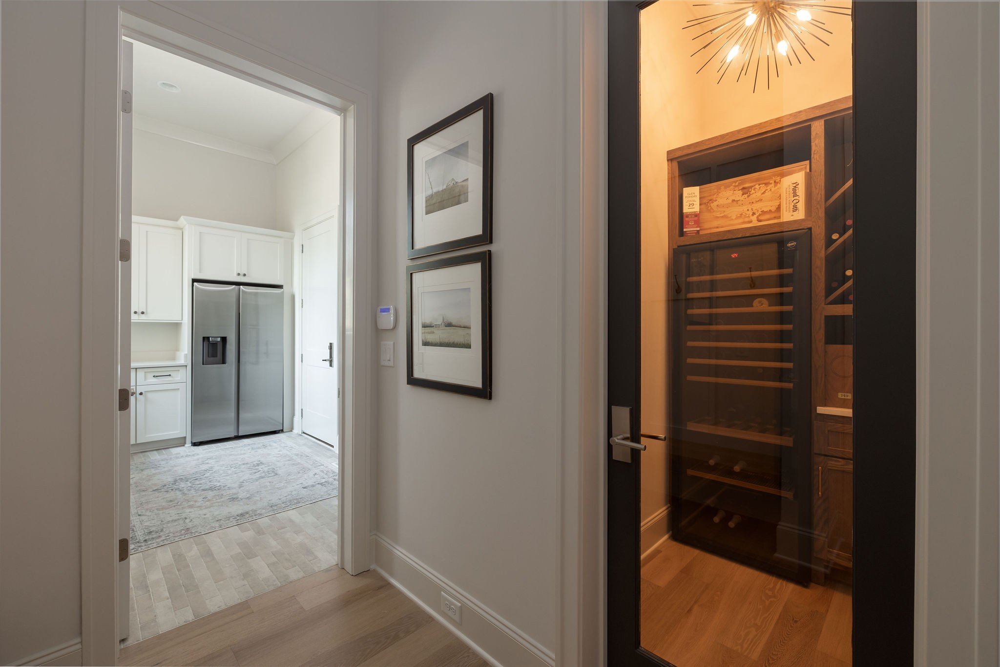 Wine cellar with wood racks and glass door beside a bright pantry with white cabinetry and stainless refrigerator.