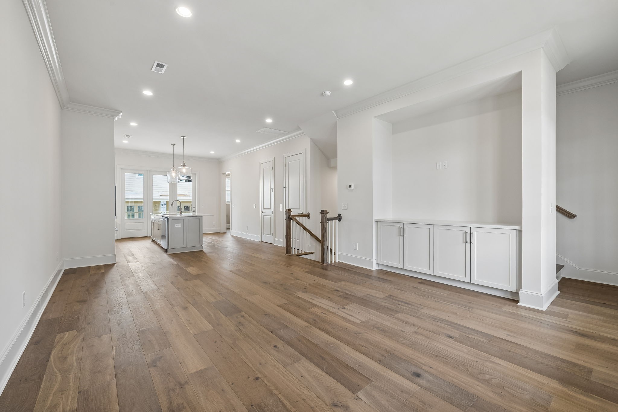 Open, vacant interior with wood floors, a kitchen island, white built-in cabinets, and a stair railing.