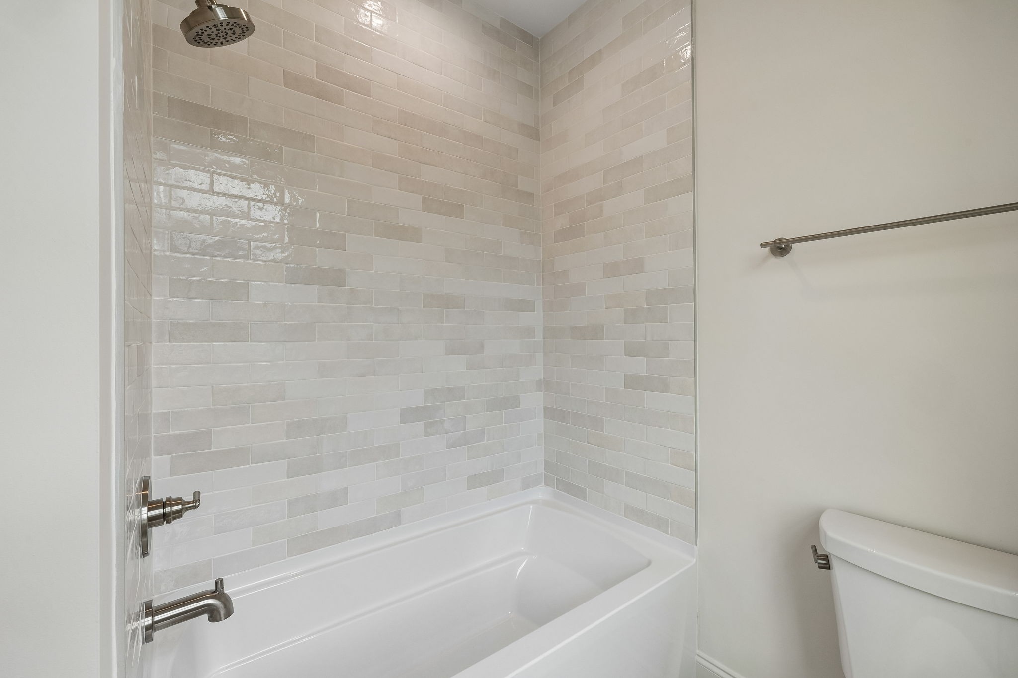 Bathroom with a white tub, light tiled shower walls, overhead showerhead, and adjacent toilet.