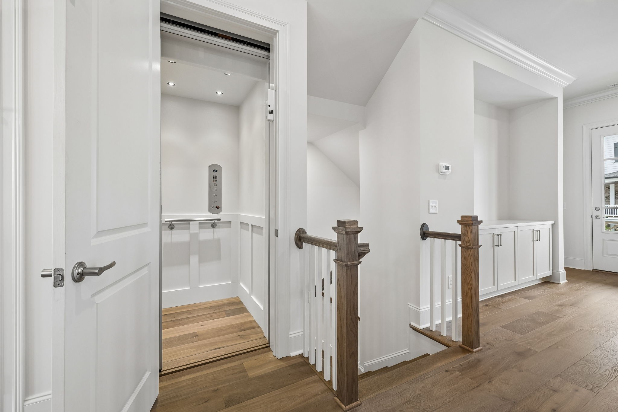 Residential landing with an open elevator, hardwood floors, and a staircase with dark wood posts.