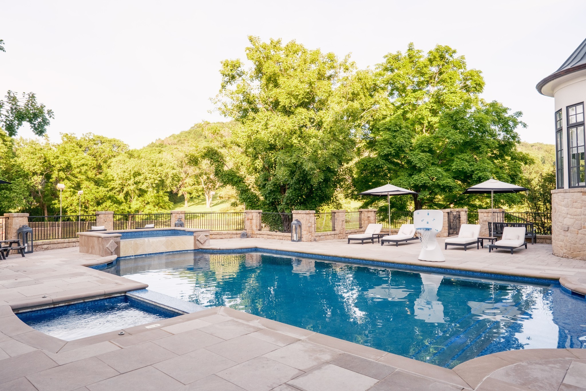 Outdoor pool and raised spa with stone patio, lounge chairs, umbrellas, and wooded hills beyond.