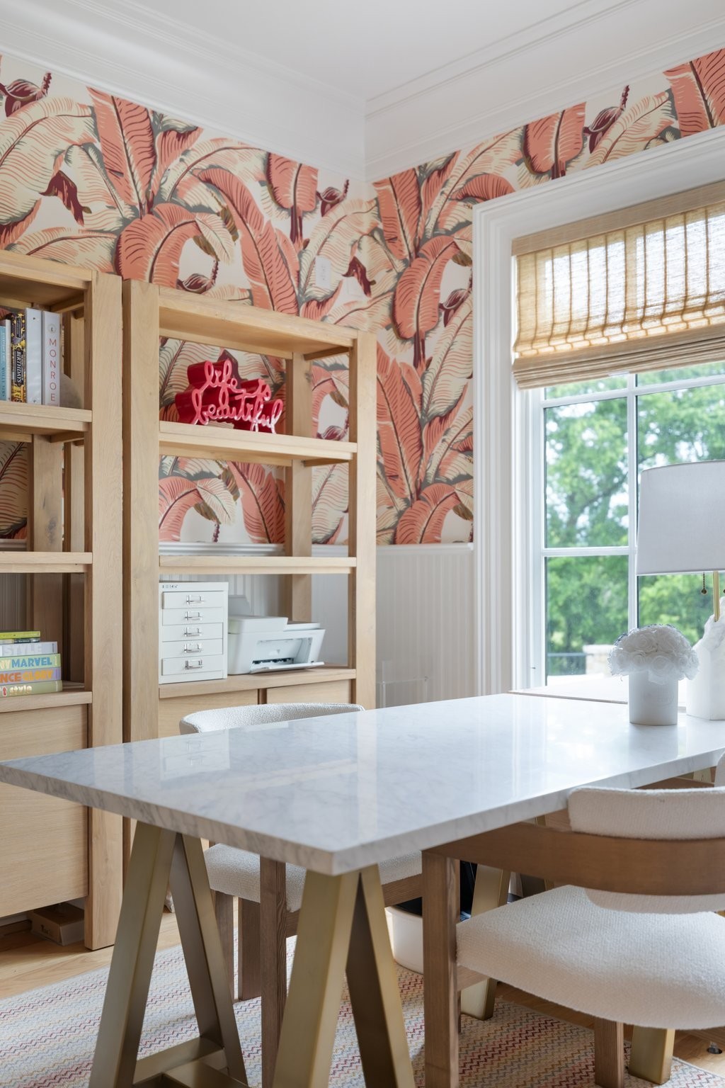 Home office with a white stone worktable, pale wood shelving, and coral tropical-leaf wallpaper.