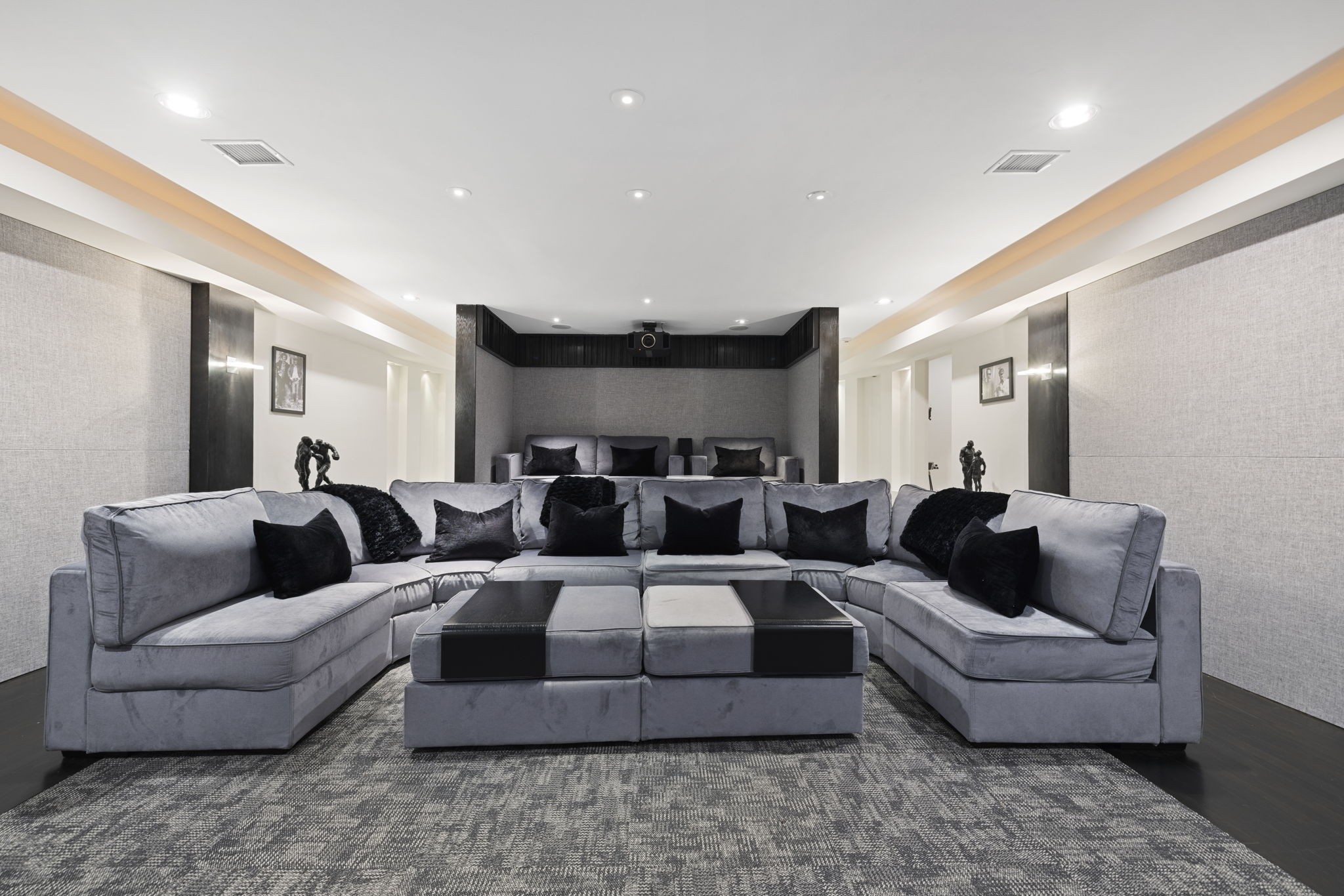 Home theater with a broad gray sectional, black pillows, acoustic wall panels, and ceiling-mounted projector.