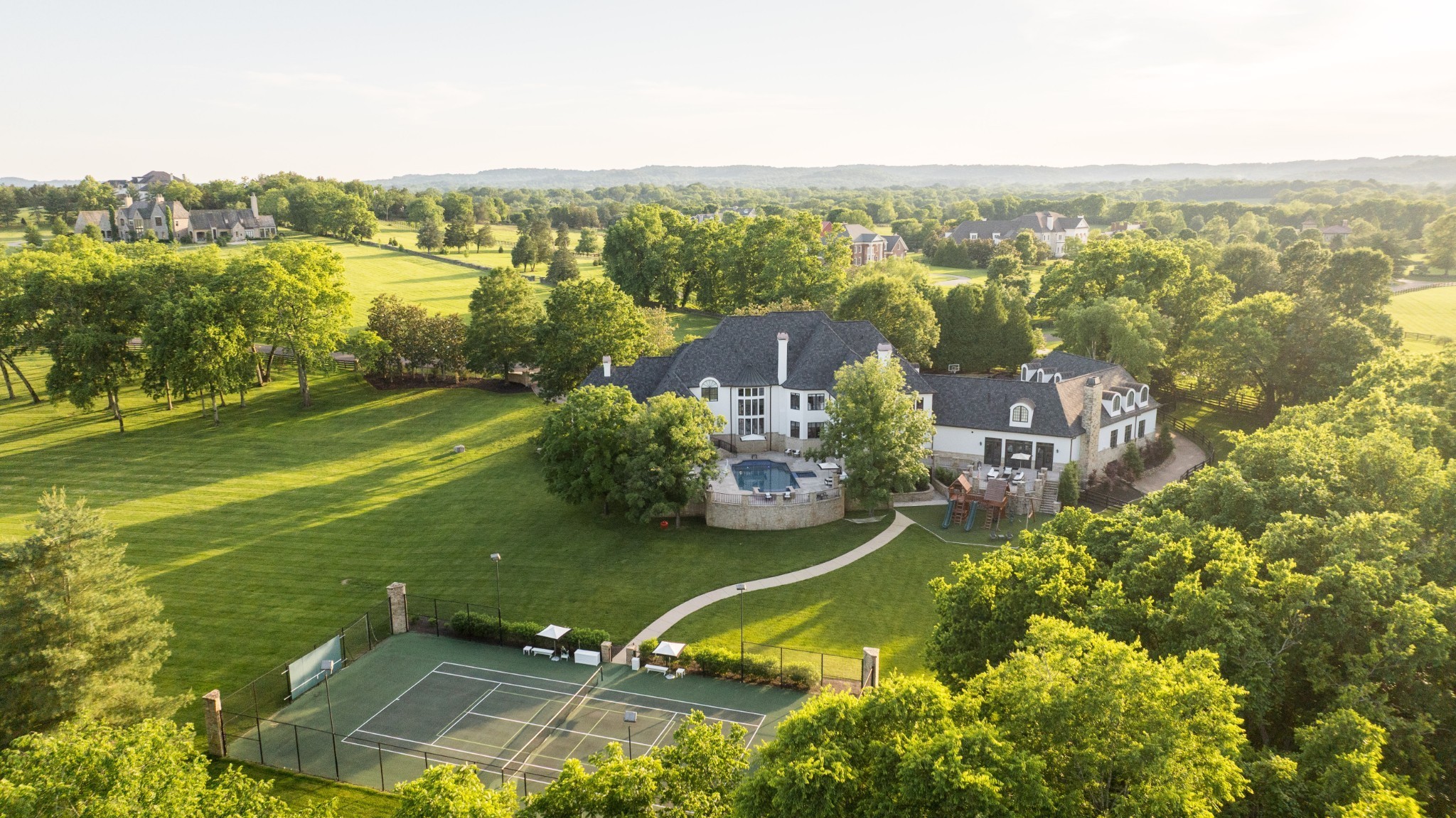 Aerial view of a large estate with a pool, tennis court, and expansive tree-lined grounds.