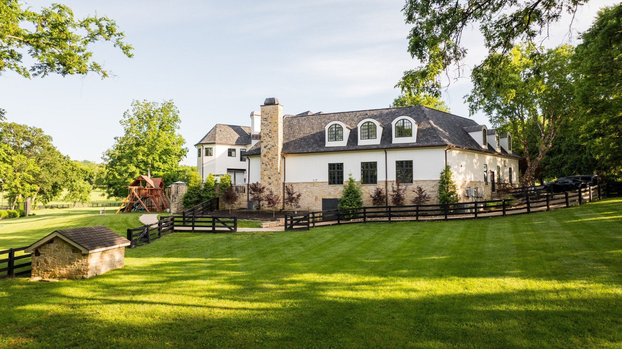 Large white and stone house with arched dormers, a fenced sloping lawn, and a wooden playset.