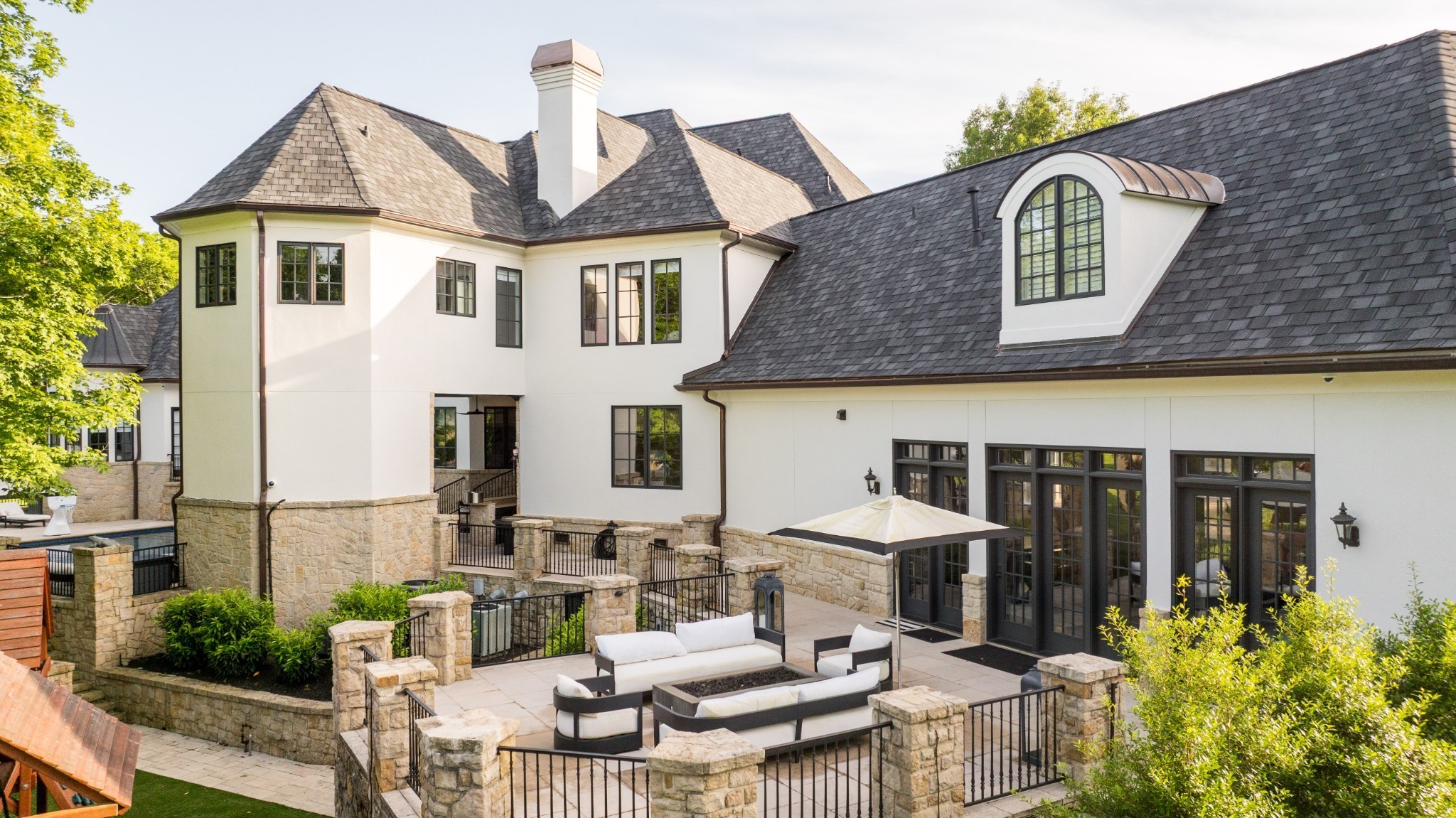 Rear of a large white house with a stone patio, outdoor seating, and a glimpse of the pool.