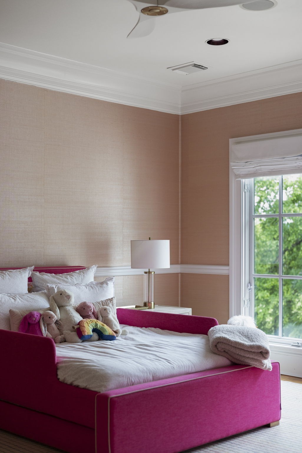 Bedroom with a bright pink bed, stuffed animals, peach walls, and a large window.