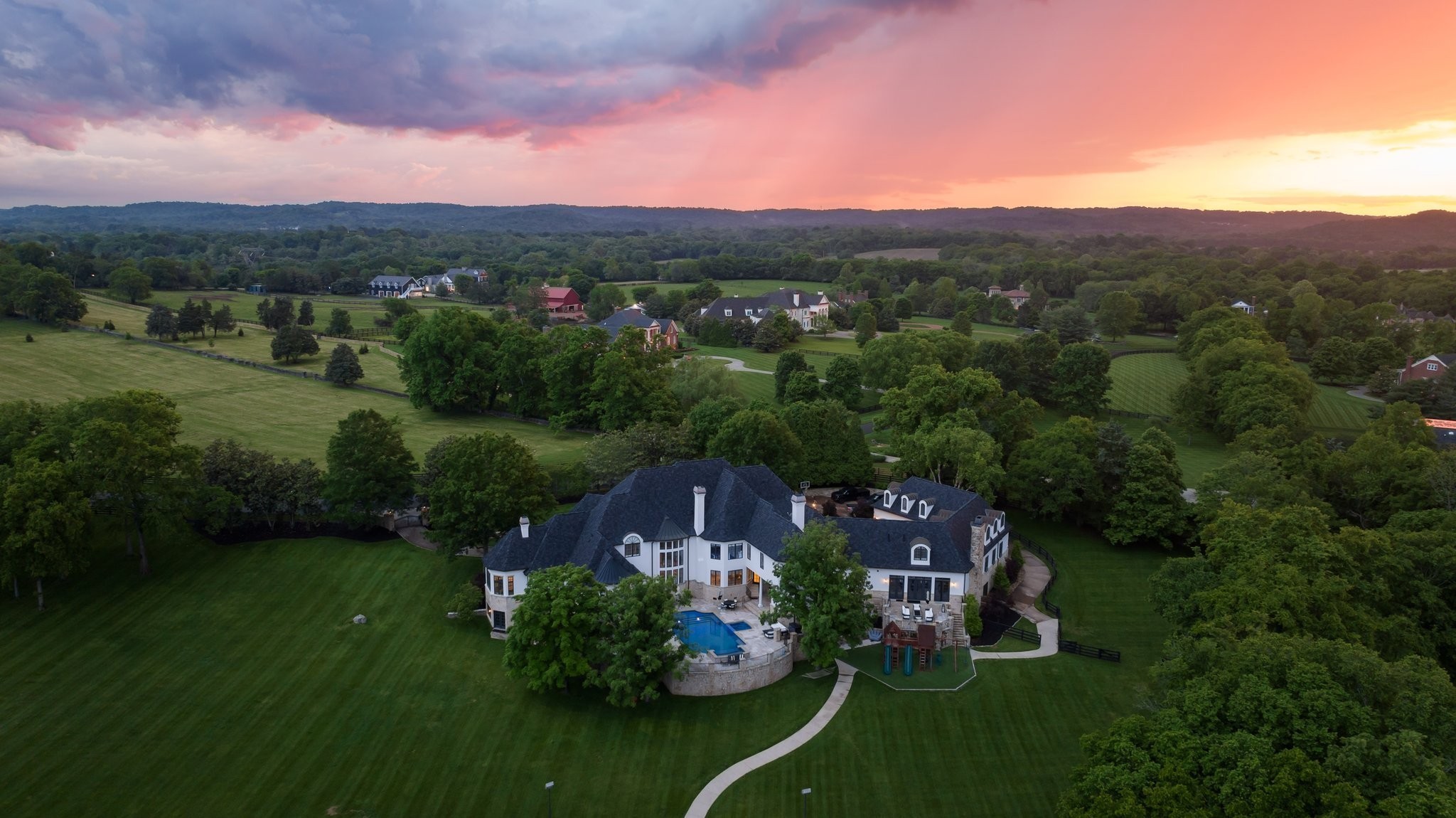 Aerial view of a large residence, swimming pool, and expansive lawns beneath a colorful sunset.